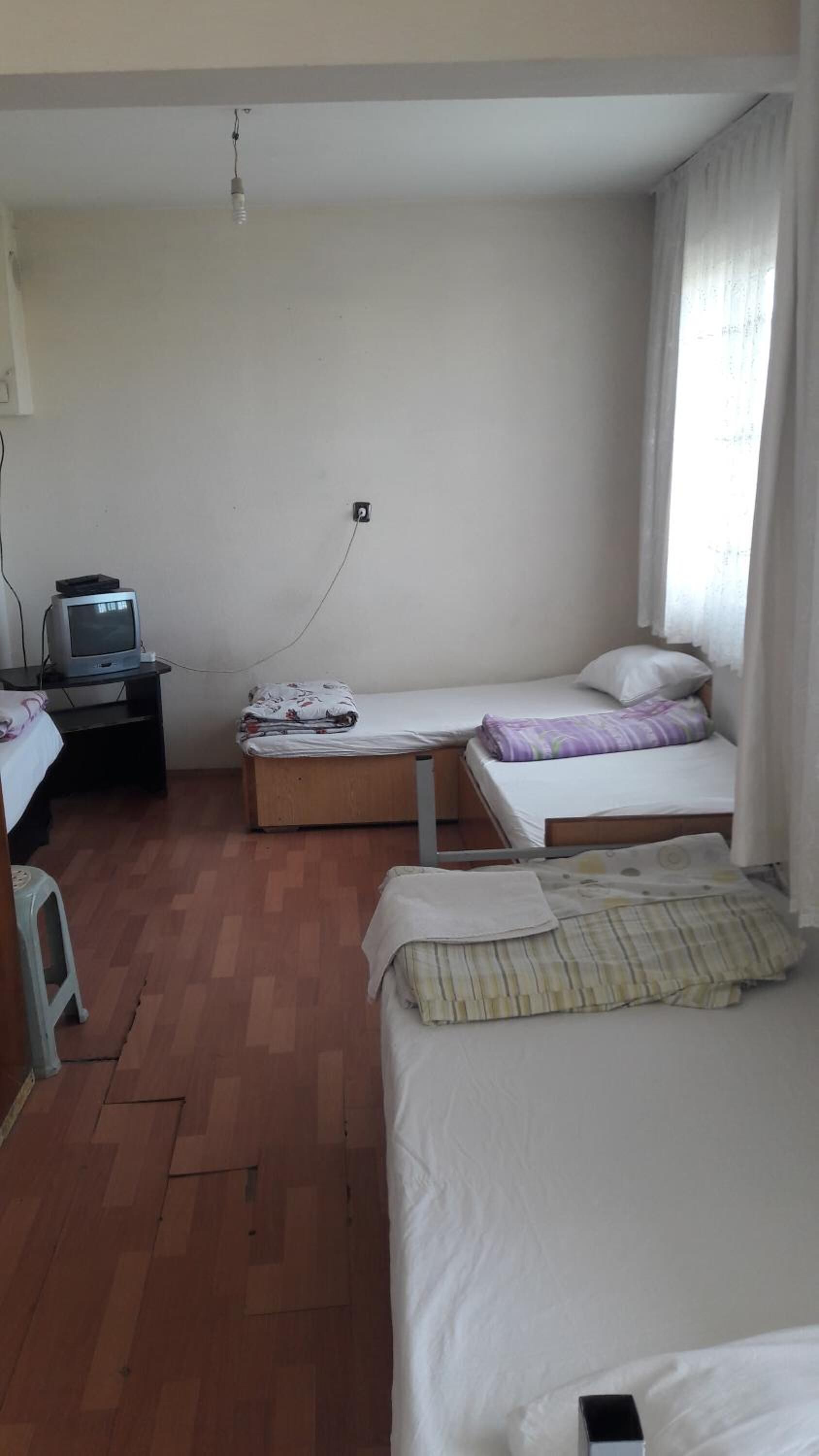 A small, sparsely furnished room with two beds, a desk with a television, and a power outlet on the wall.