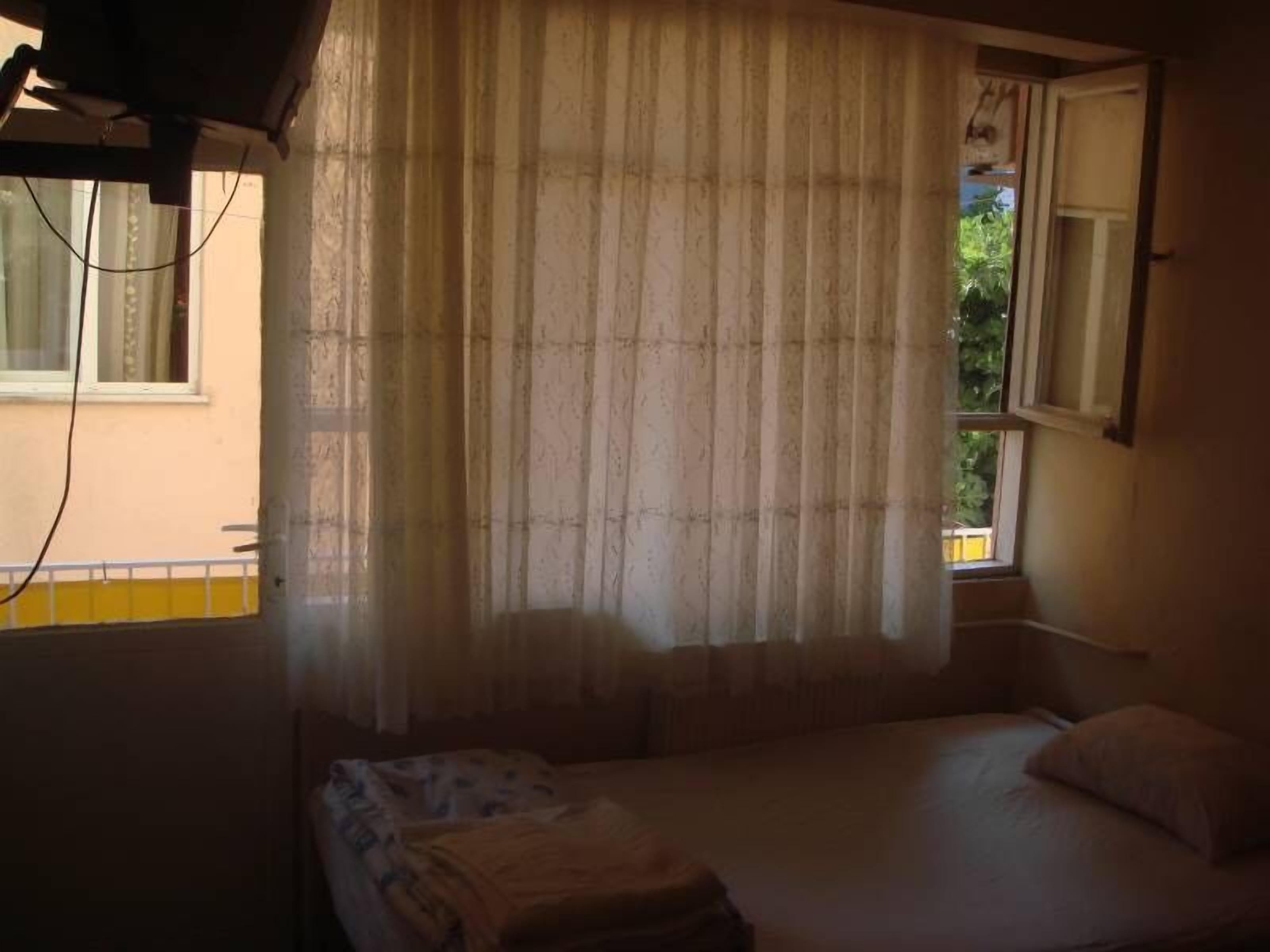 Um quarto pequeno e com pouca luz, com uma cama de solteiro, uma janela com cortinas e uma prateleira fixada na parede.