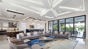 Ein modernes Wohnzimmer mit einer großen Eckcouch, einem Couchtisch und einem gemusterten Teppich. Es gibt einen Küchenbereich mit integrierter Kochinsel und einen Essbereich, der durch eine Glaswand abgetrennt ist.