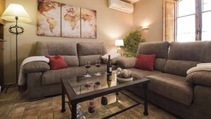 Ein Wohnzimmer mit einer Sofagarnitur, einem Couchtisch mit Weingläsern und einer Flasche, einem Wandbild mit einer Weltkarte, einer Klimaanlage und einem Fenster mit Tageslicht.