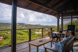 A covered outdoor area with a table and chairs, a grill, and a view of a green landscape.