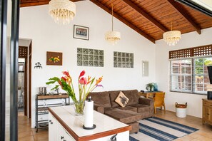 A living room with a brown sofa, a coffee table, and a vase of flowers. There are three pendant lights, a wooden ceiling, and a window with blinds. - Ocean-view 1BR Orchid Cottage in Cap Estate with private pool, patio & A/C. (Cap Estate)