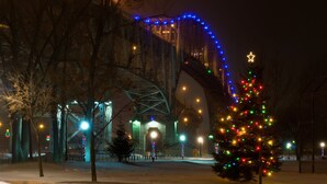 A snow-covered park with a bridge, decorated with blue and white lights, and a Christmas tree with multicolored lights.