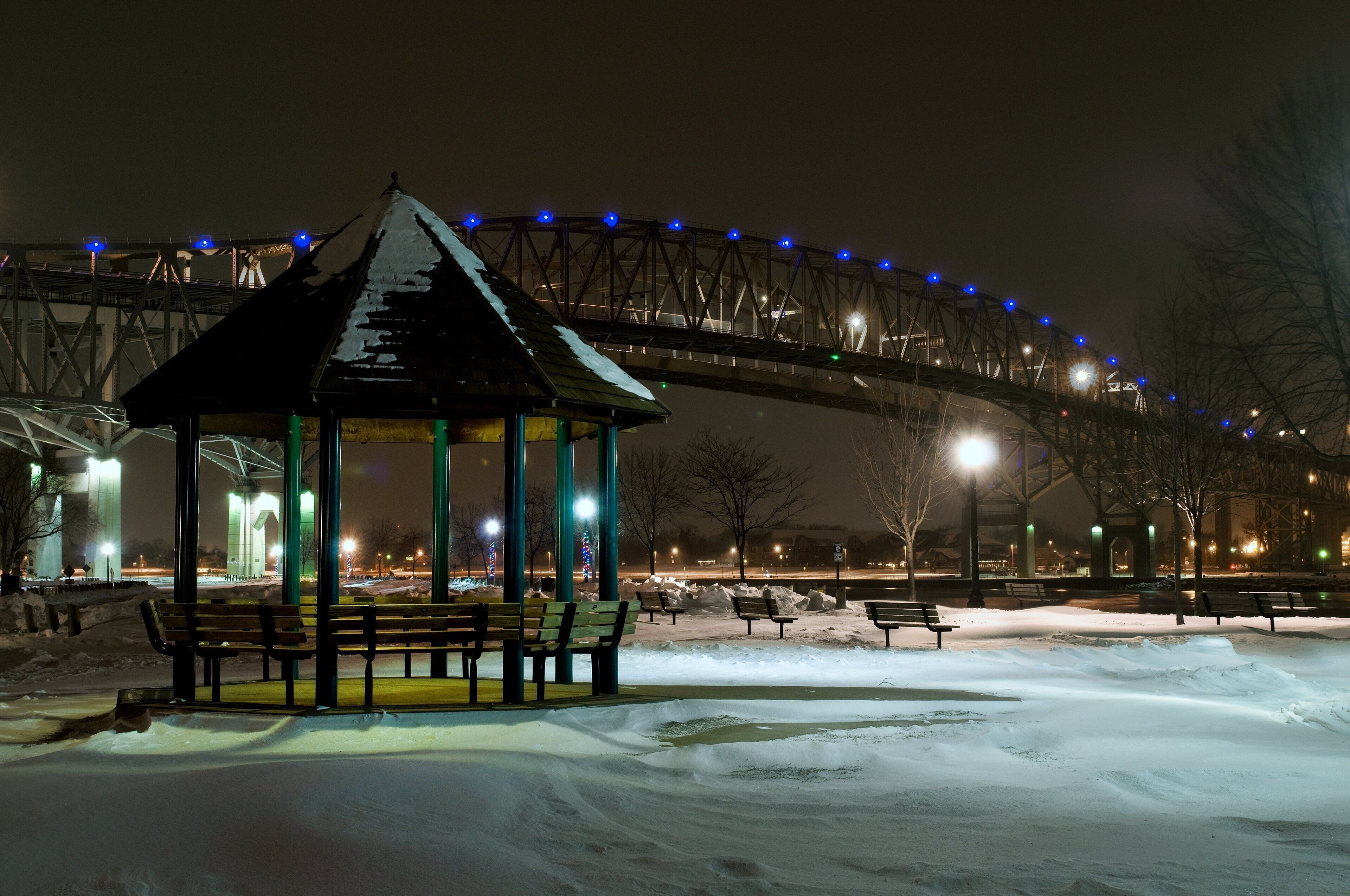 Un jardín cubierto de nieve con bancos, en un parque nocturno iluminado por farolas y bajo un puente con luces azules.