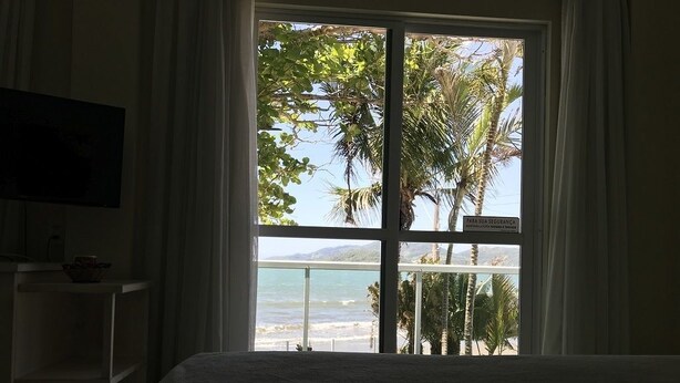 Habitación de hotel con cama, televisor sobre un mueble bajo y ventana con vistas a la playa y palmeras.