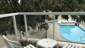 Un balcón con sillas de plástico blanco y una pequeña mesa, con vistas a una piscina y un área vallada con sillas de descanso.
