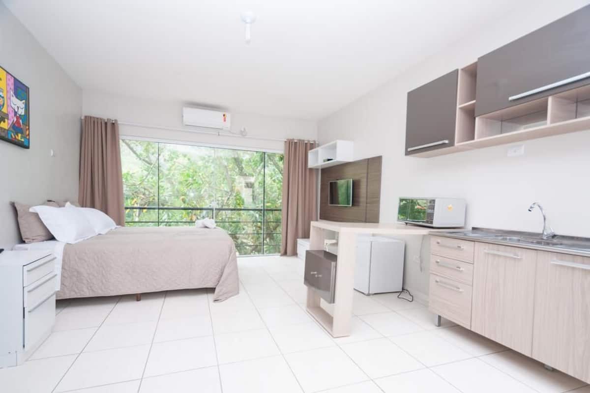 A modern hotel room with a bed, kitchenette, and a large window with curtains.