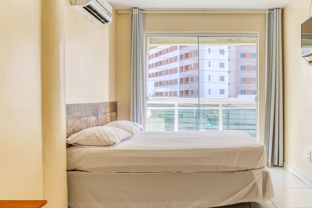 Une chambre d’hôtel équipée d’un lit, d’un appareil de climatisation, d’une fenêtre avec des rideaux et offrant une vue sur un bâtiment situé à l’extérieur.