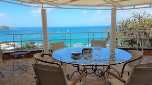 Une table ronde en verre, entourée de chaises, placée sur une terrasse offrant une vue sur la mer et les bateaux.