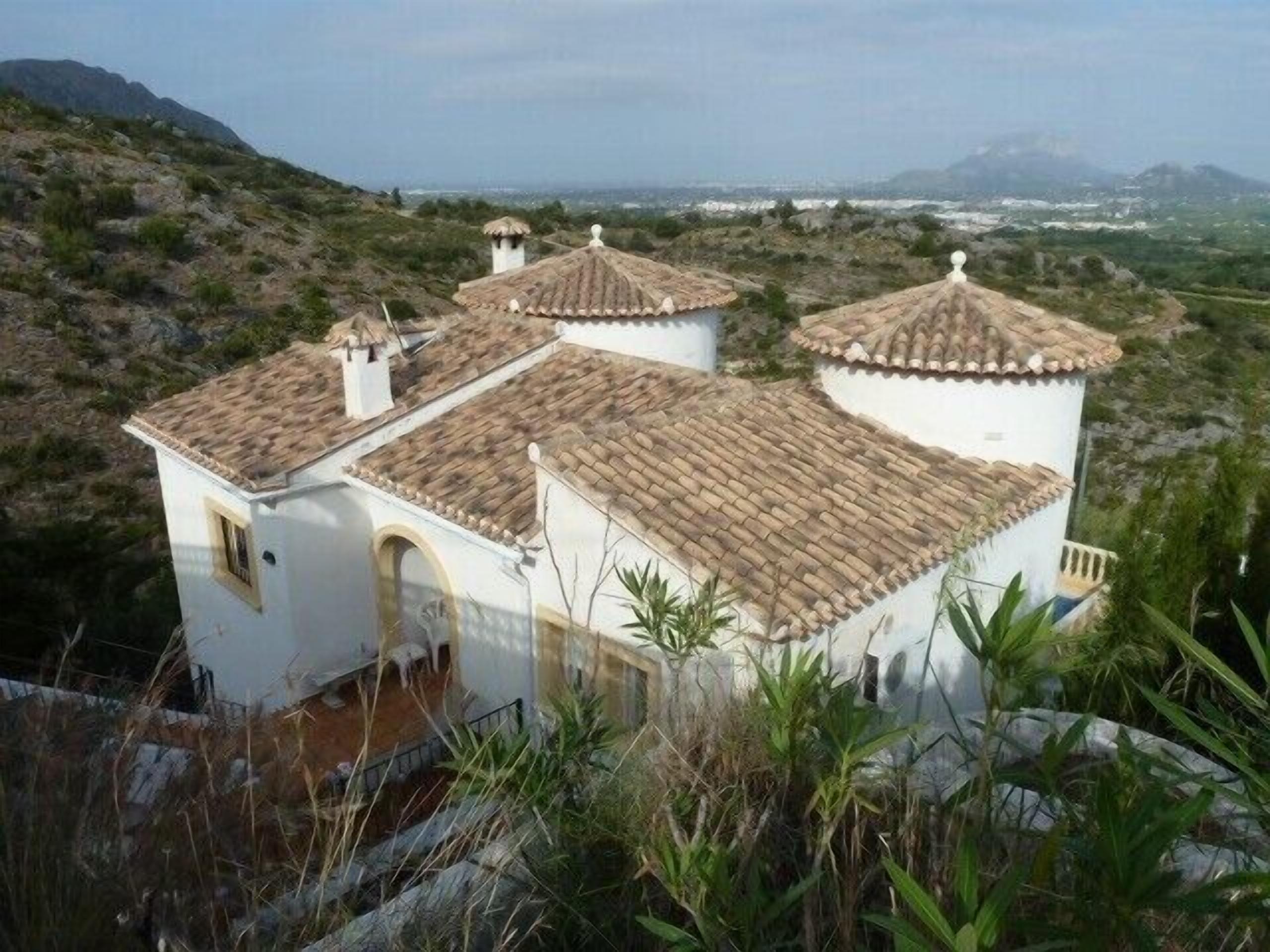 A white building with terracotta roofs, surrounded by greenery and mountains in the background.