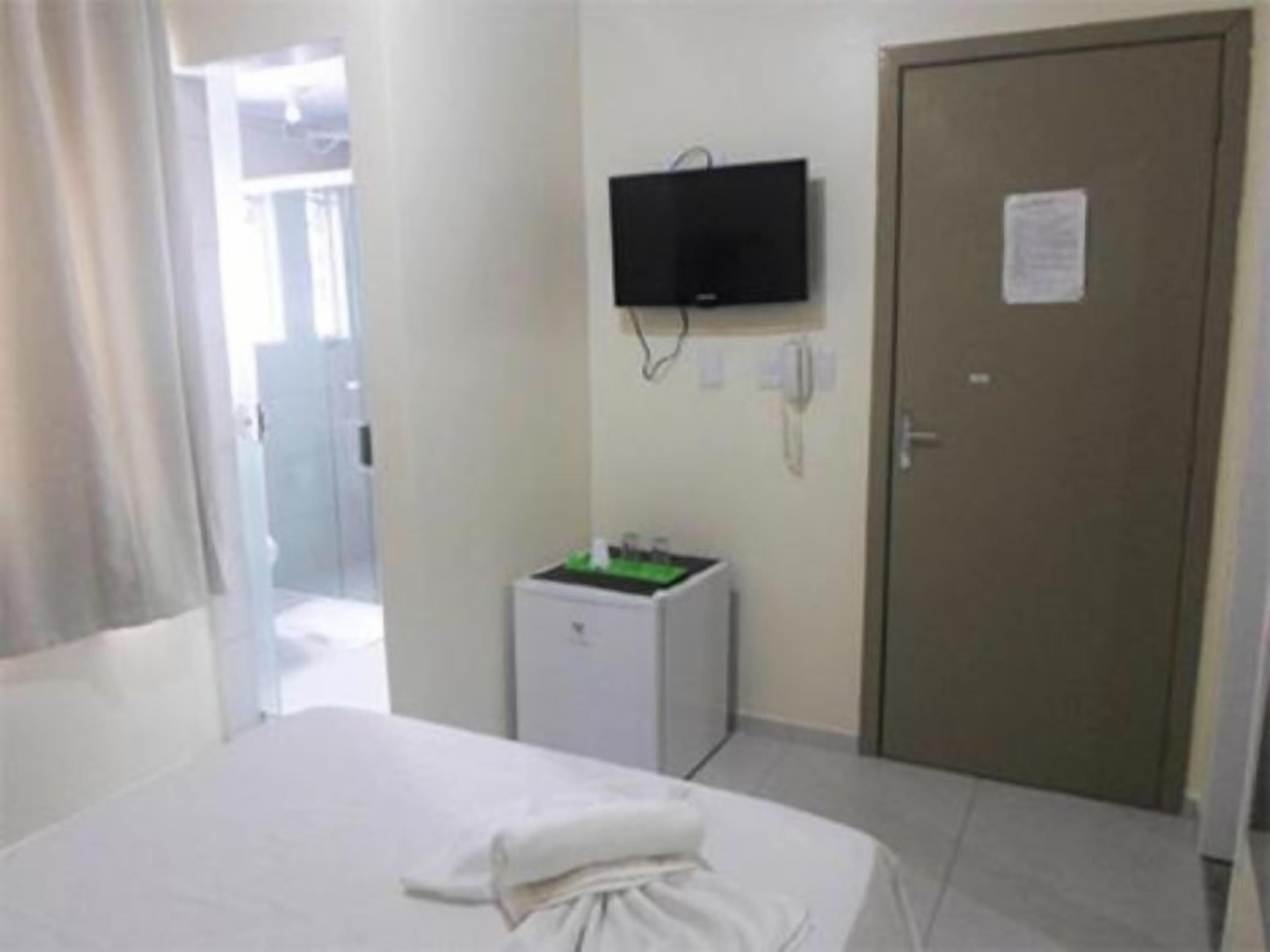 Una habitación de hotel con una cama, un televisor montado en la pared, un escritorio pequeño con un objeto verde encima y una puerta con un letrero.