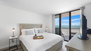 A hotel room with a large bed, a bedside table with a lamp, a tray with a bottle and glasses, and a balcony with a view of the ocean. - Modern 11th Floor Beachfront Condo With Views/pool (Marco Island)
