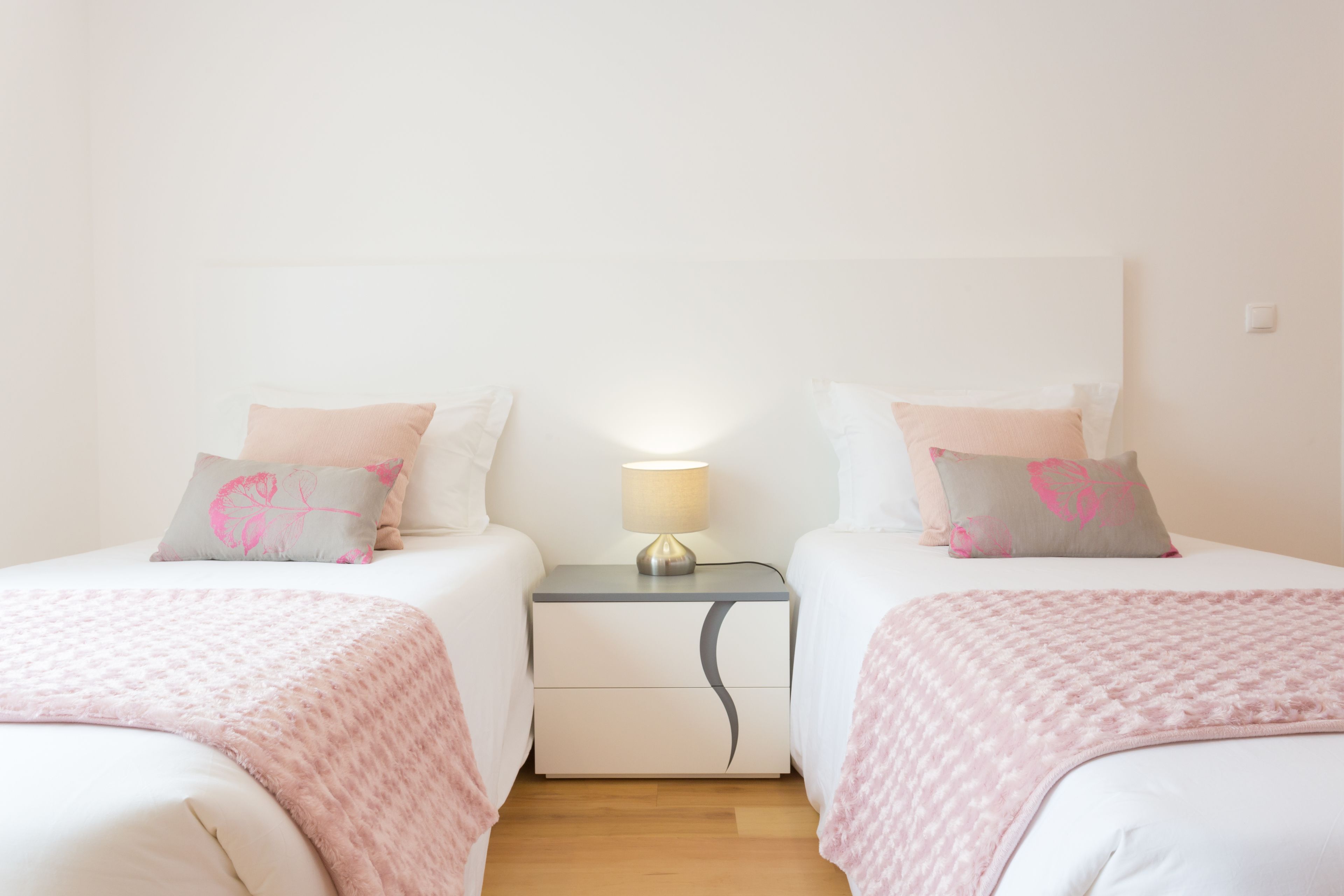 Camera matrimoniale con due letti, ciascuno con coperta rosa e cuscini abbinati, un comodino con lampada e pavimento in legno.