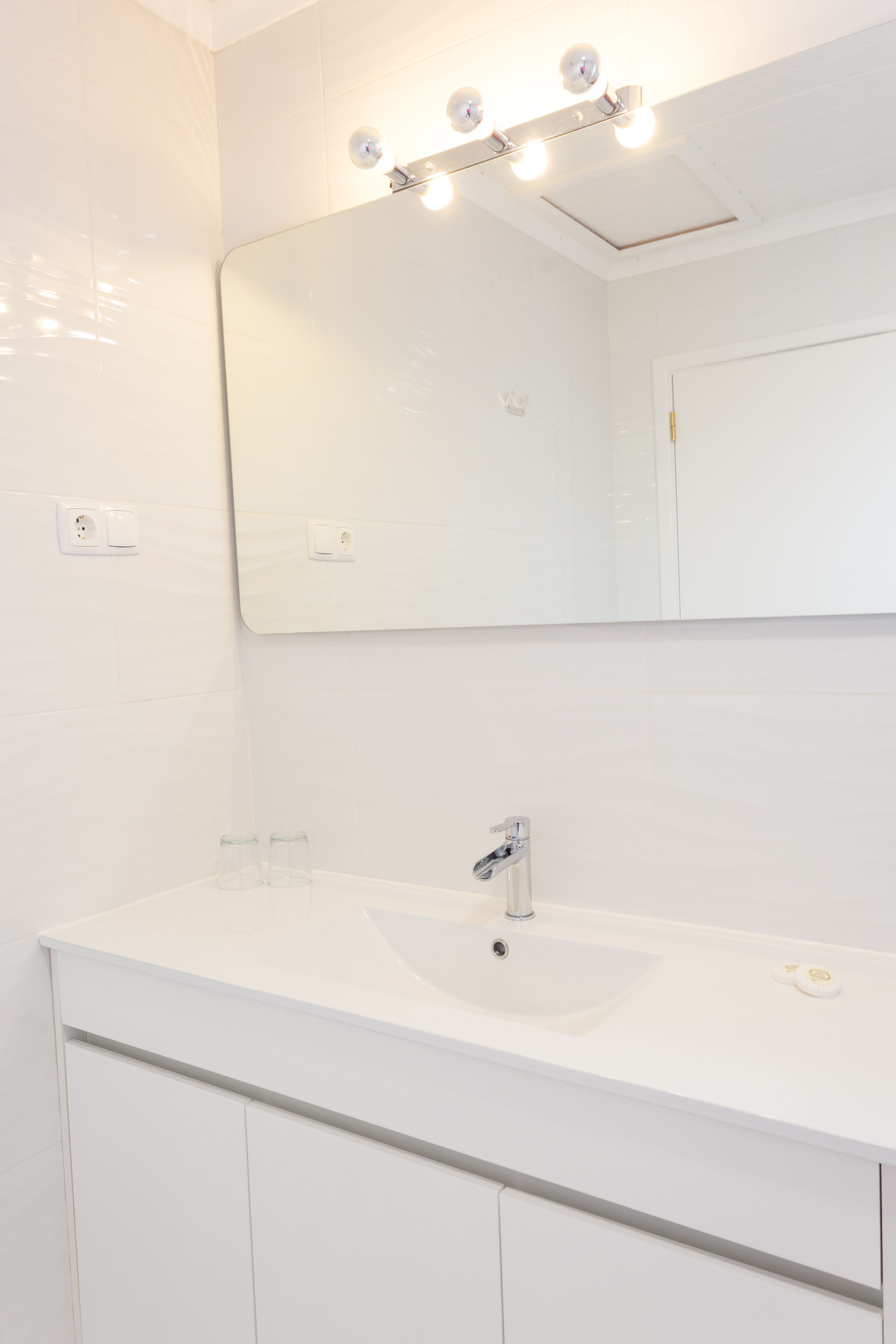 Un baño con lavamanos blanco, espejo e iluminación sobre el espejo.