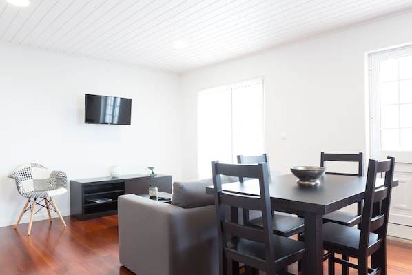 A modern living room with a dark wooden dining table, grey sofa, and a black TV mounted on the wall. - 35th Atlantic View (Lajes do Pico)