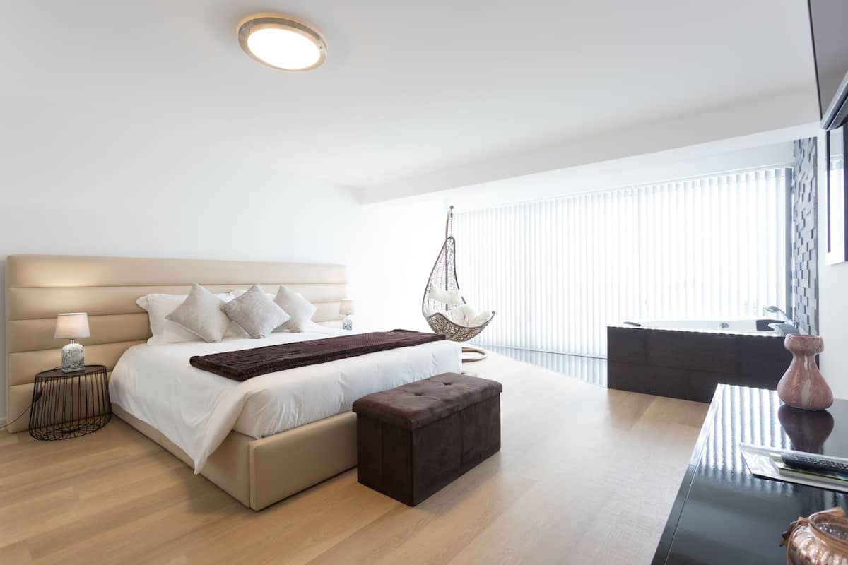 A modern bedroom with a large bed, bedside tables, a hanging chair, a small table, and a view of a balcony.