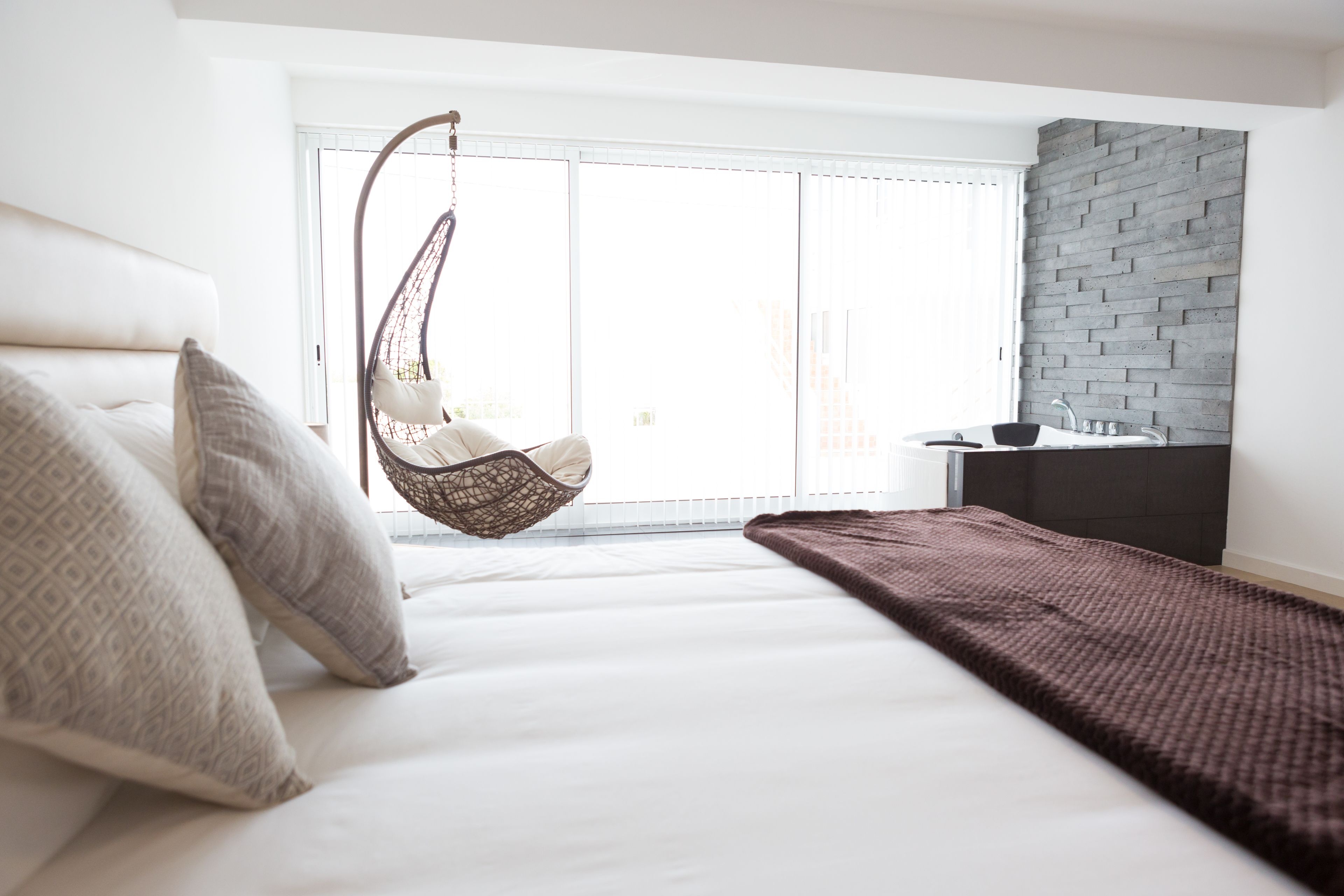Un dormitorio moderno con una silla colgante, cama con almohadas estampadas, cabecero oscuro y vistas de la ciudad a través de amplias ventanas.
