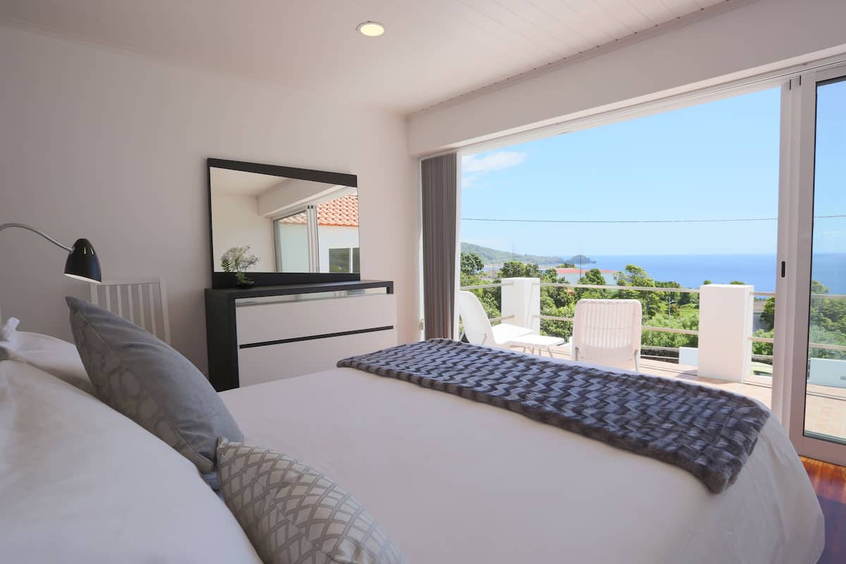A bedroom with a bed, a dresser, a mirror, and a balcony with a view of the sea.
