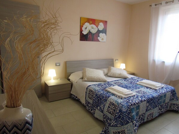 A hotel room with a bed, nightstands, a vase with dried plants, a painting, and a window with curtains.