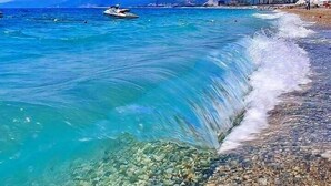 Una playa con aguas turquesas cristalinas, orilla pedregosa y montañas al fondo.