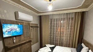 Habitación de hotel con cama, televisor montado en la pared, lámpara de techo y ventana con cortinas.
