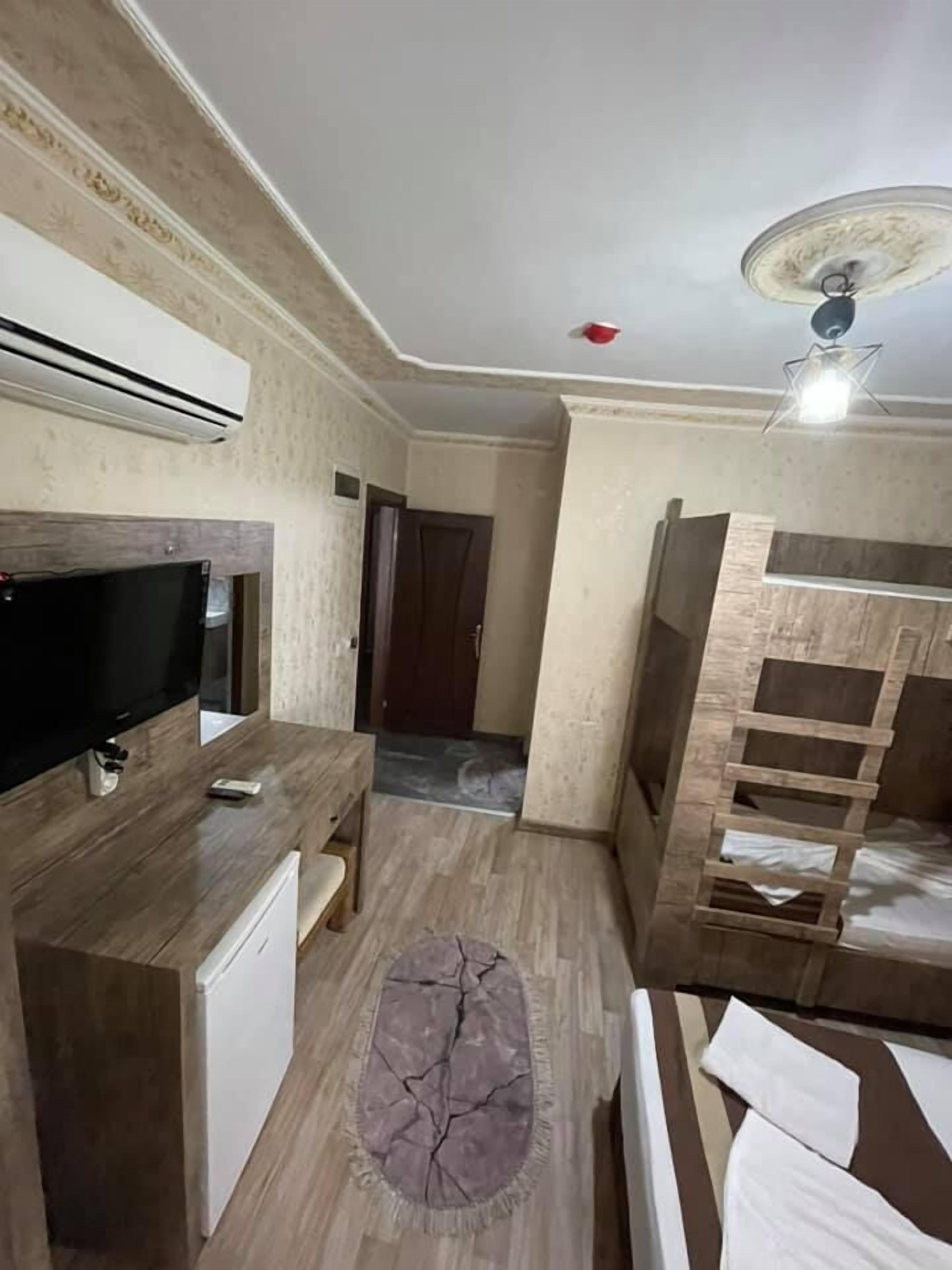 A room with a TV, a small fridge, a wooden desk, a bed with a ladder, and a ceiling fan.
