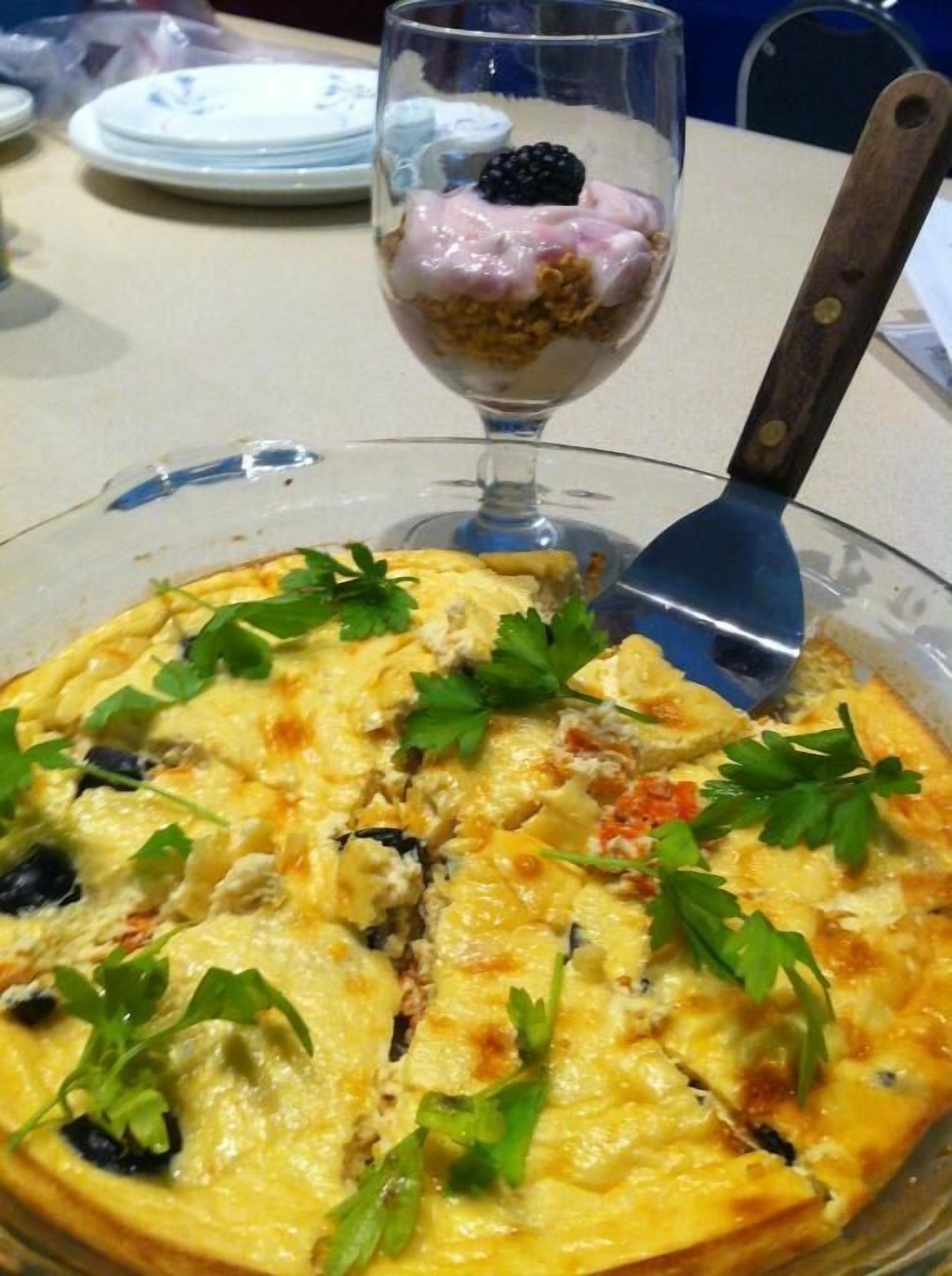 A quiche with parsley garnish, a glass of dessert with berries, and a knife on a table.