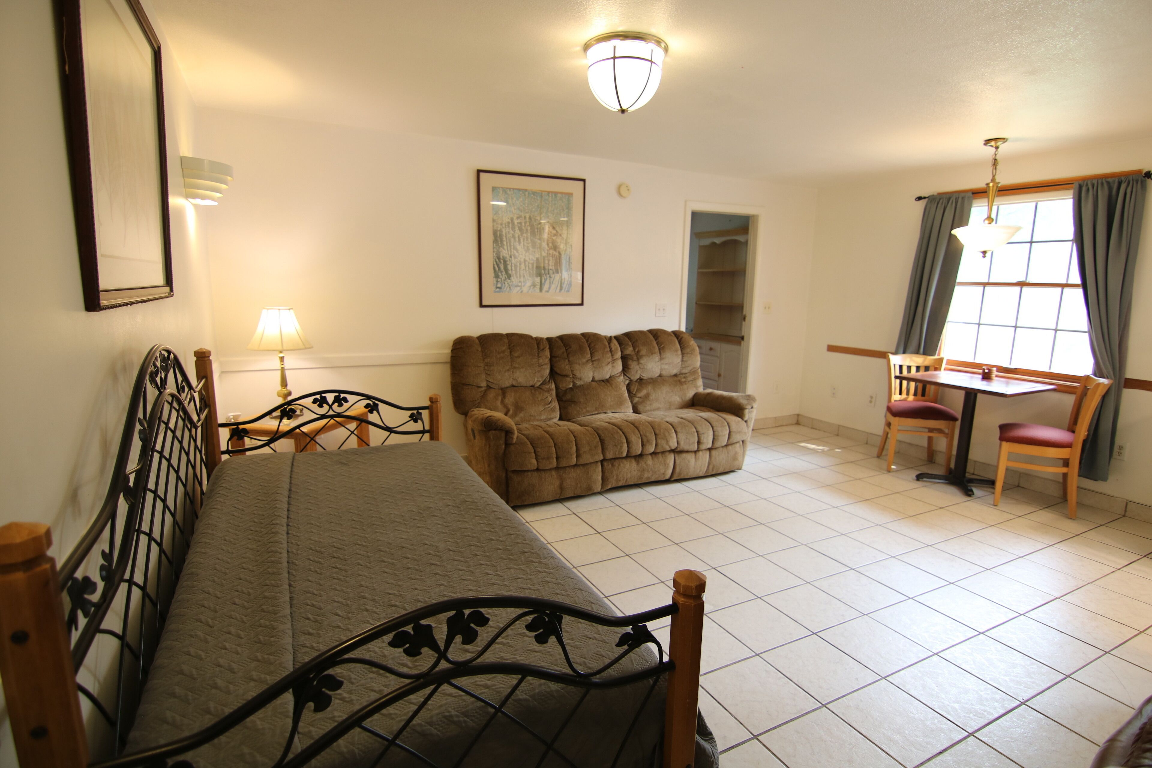 A room with a bed, a sofa, a dining table with chairs, a window with curtains, and a wall-mounted lamp.