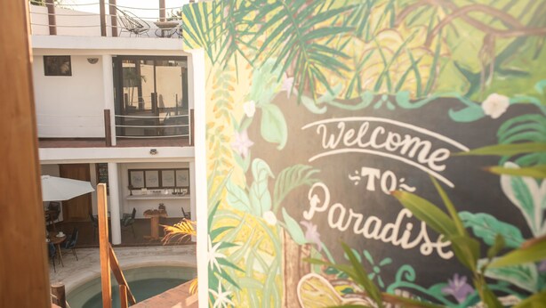A pool area with a 'Welcome to Paradise' sign, indoor seating, and a view of the upper floor. - Hotel Ade (Bacalar)