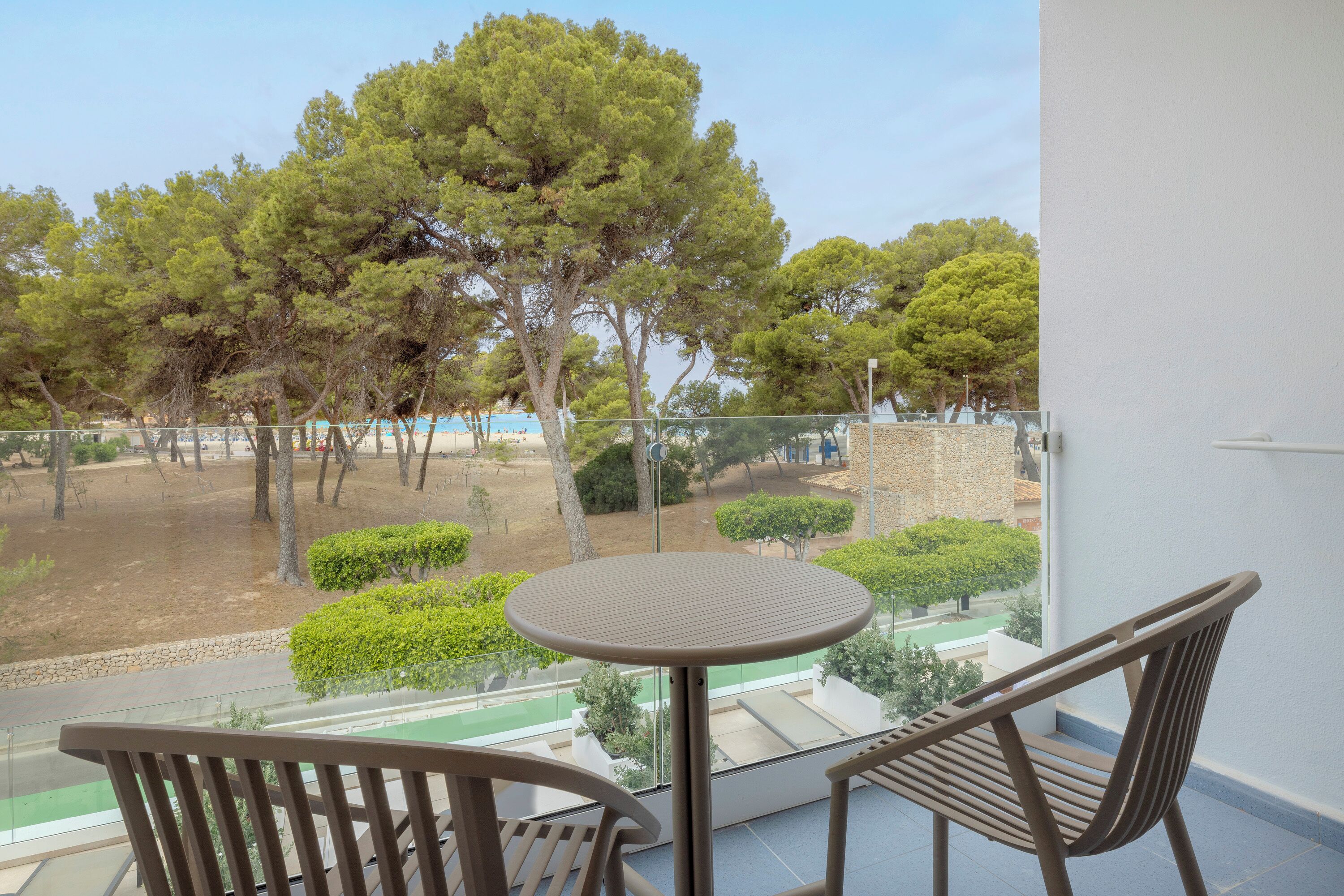 A balcony with a round table and two chairs, overlooking a landscaped area with trees and a distant view of a building.
