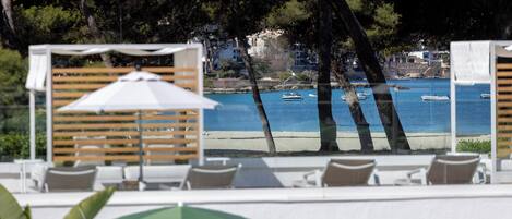 A poolside area with lounge chairs and umbrellas, overlooking a beach with boats and buildings in the background.
