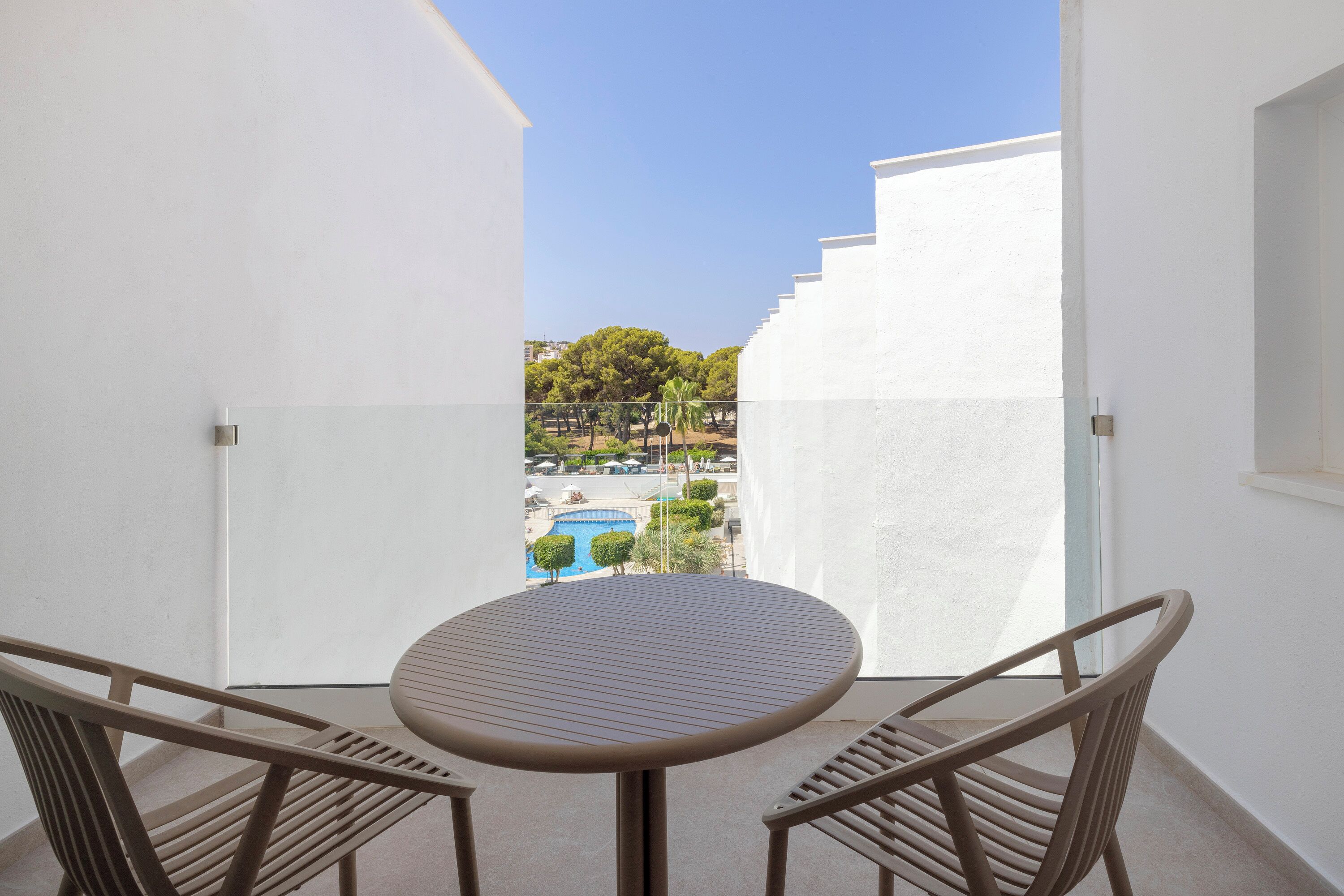 A balcony with a round table and two chairs, overlooking a swimming pool and greenery.
