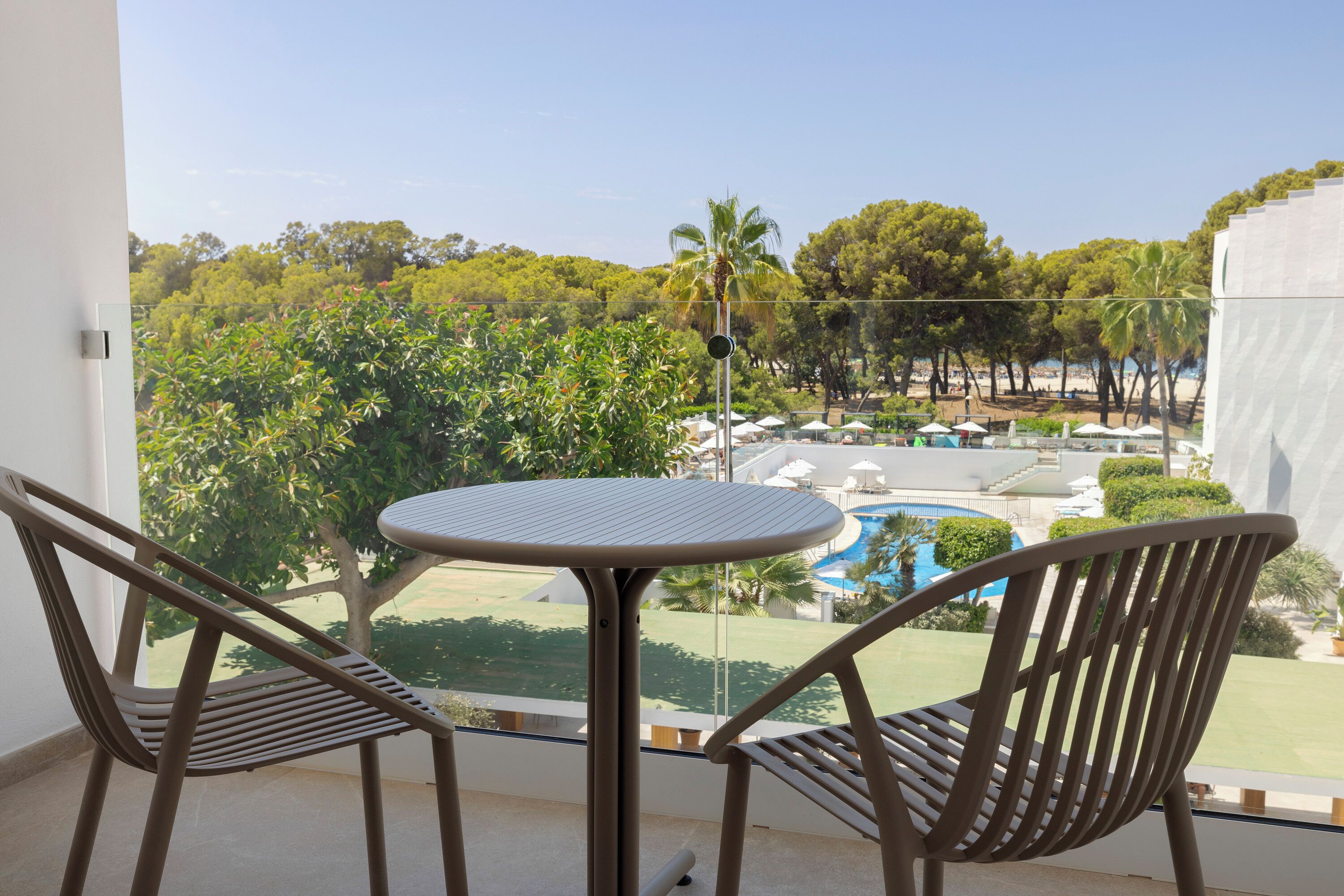 A balcony with two chairs and a table, overlooking a pool and lush greenery.