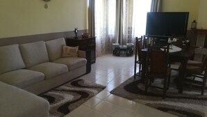 A living room with a sofa, a television, a dining table with chairs, and a decorative clock on the wall. - HOLLY - Quiet guest house with seaview and a pool (Balchik)