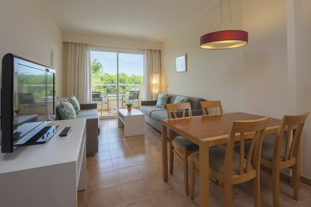 A modern living room with a flat-screen TV, a sofa, a dining table with chairs, and a balcony with a view of greenery.