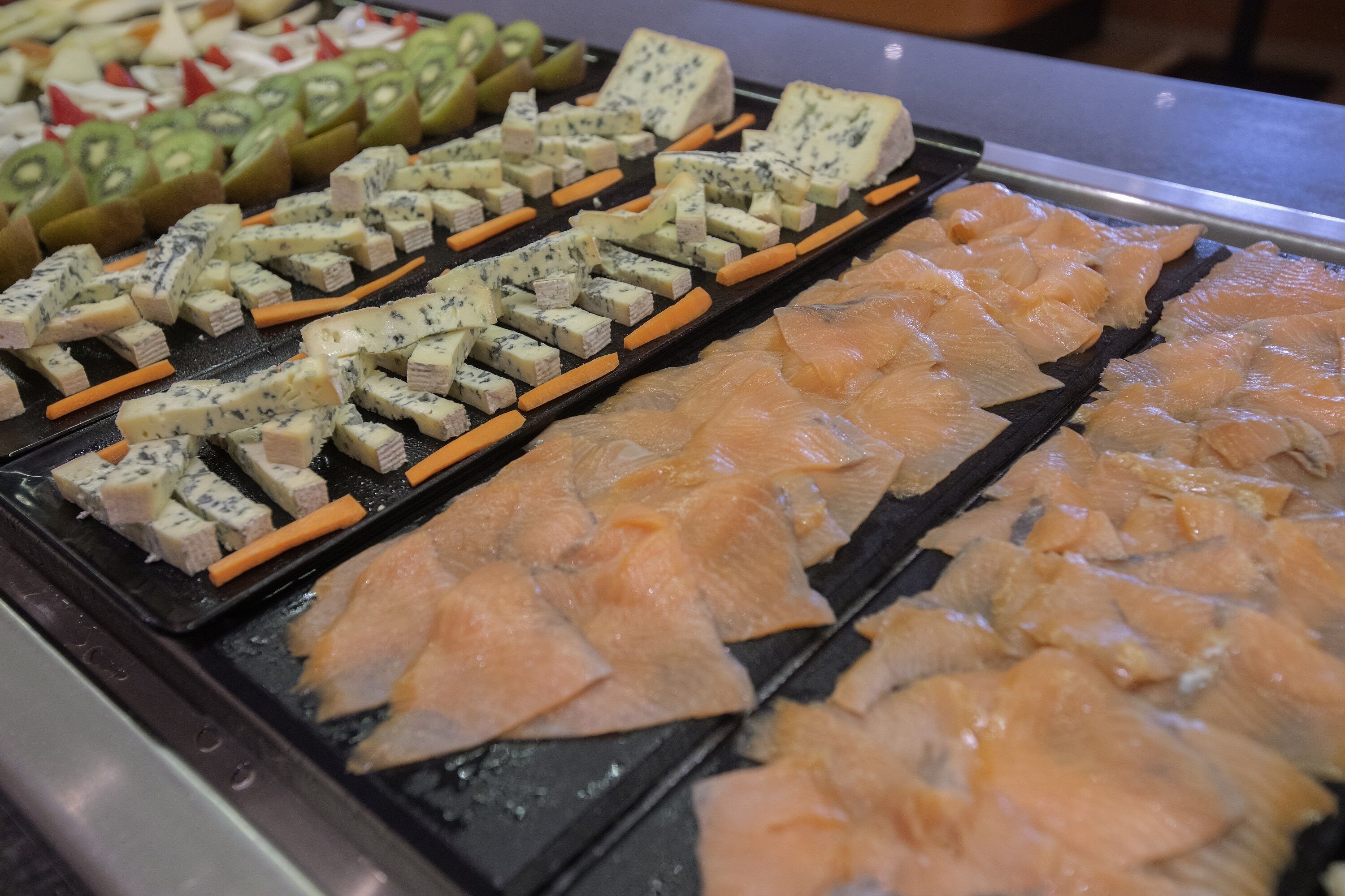 A display of various food items including sliced kiwi, blue cheese, and salmon arranged on trays.