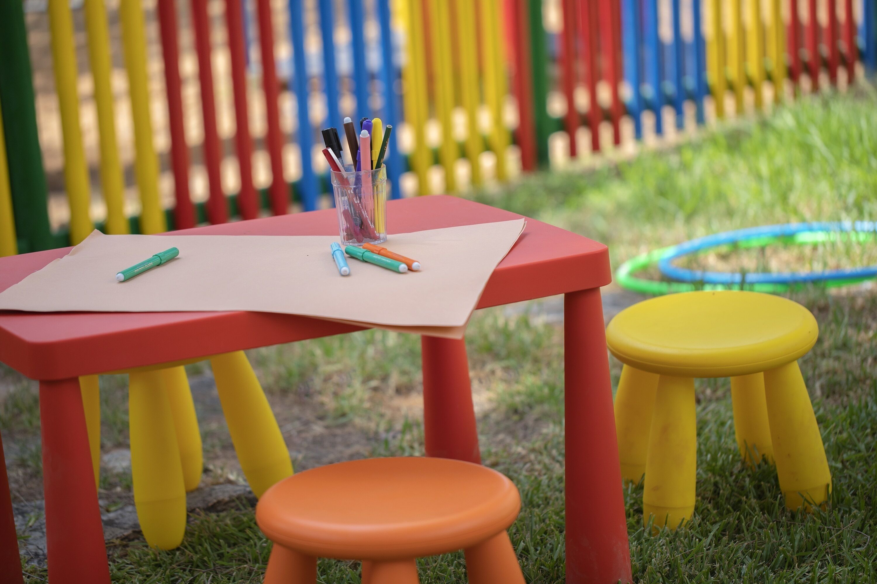 A red table with a white sheet of paper, two stools (one orange, one yellow), and a container with pens.