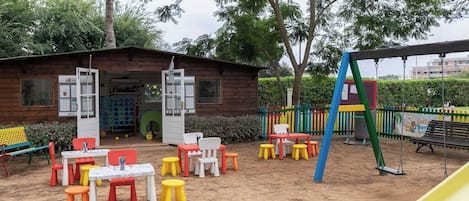A playground with colorful tables and chairs, a slide, and a wooden building with open doors.