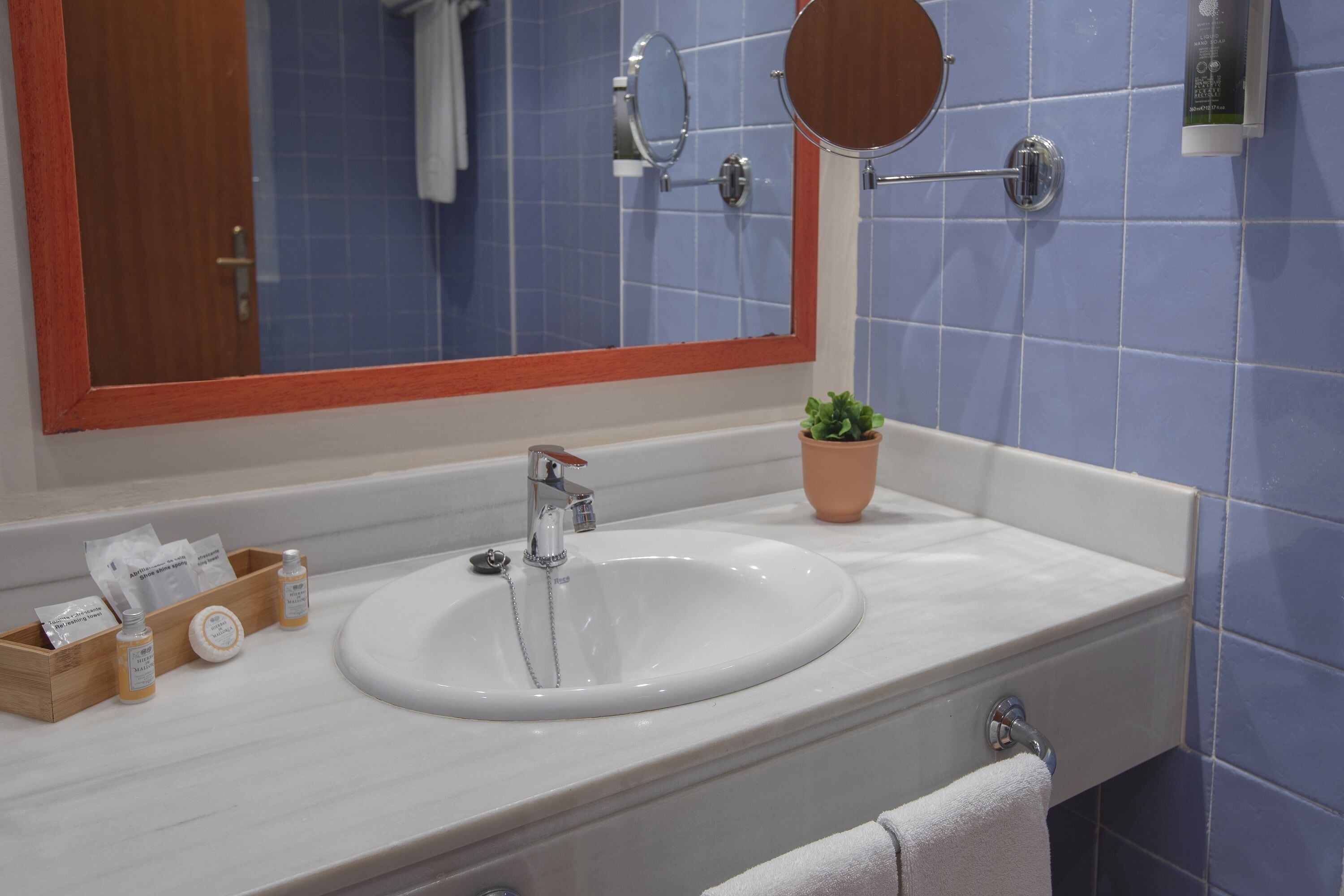 A bathroom with a white sink, a wooden tray with toiletries, a small potted plant, and a mirror with a wooden frame.