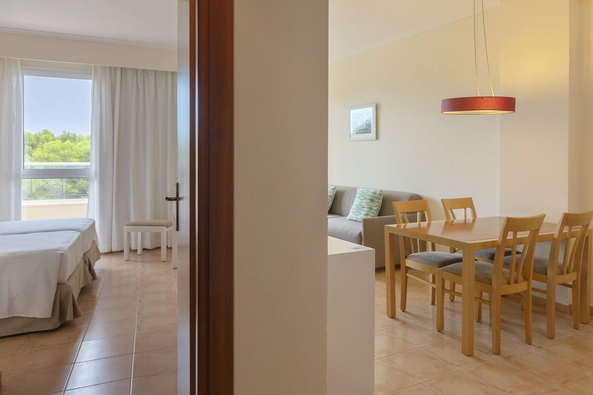 A hotel room with a balcony, a dining area with wooden chairs, a sofa, and a table.