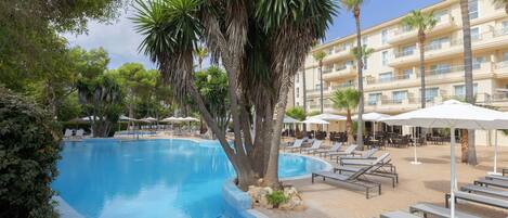 A swimming pool with lounge chairs and umbrellas, surrounded by palm trees and a building with balconies.