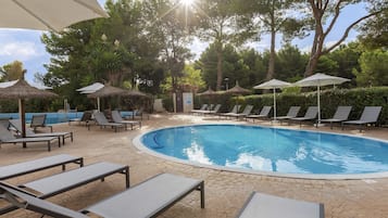 Un área de piscina con sillones de descanso y sombrillas, rodeada de árboles y un cielo despejado.