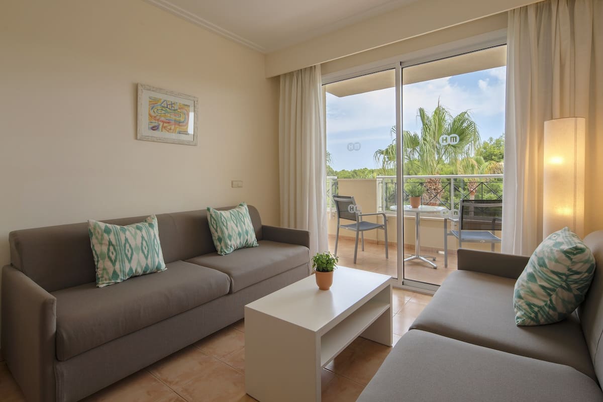 A living room with a sofa, a coffee table, and a balcony with a view of palm trees.