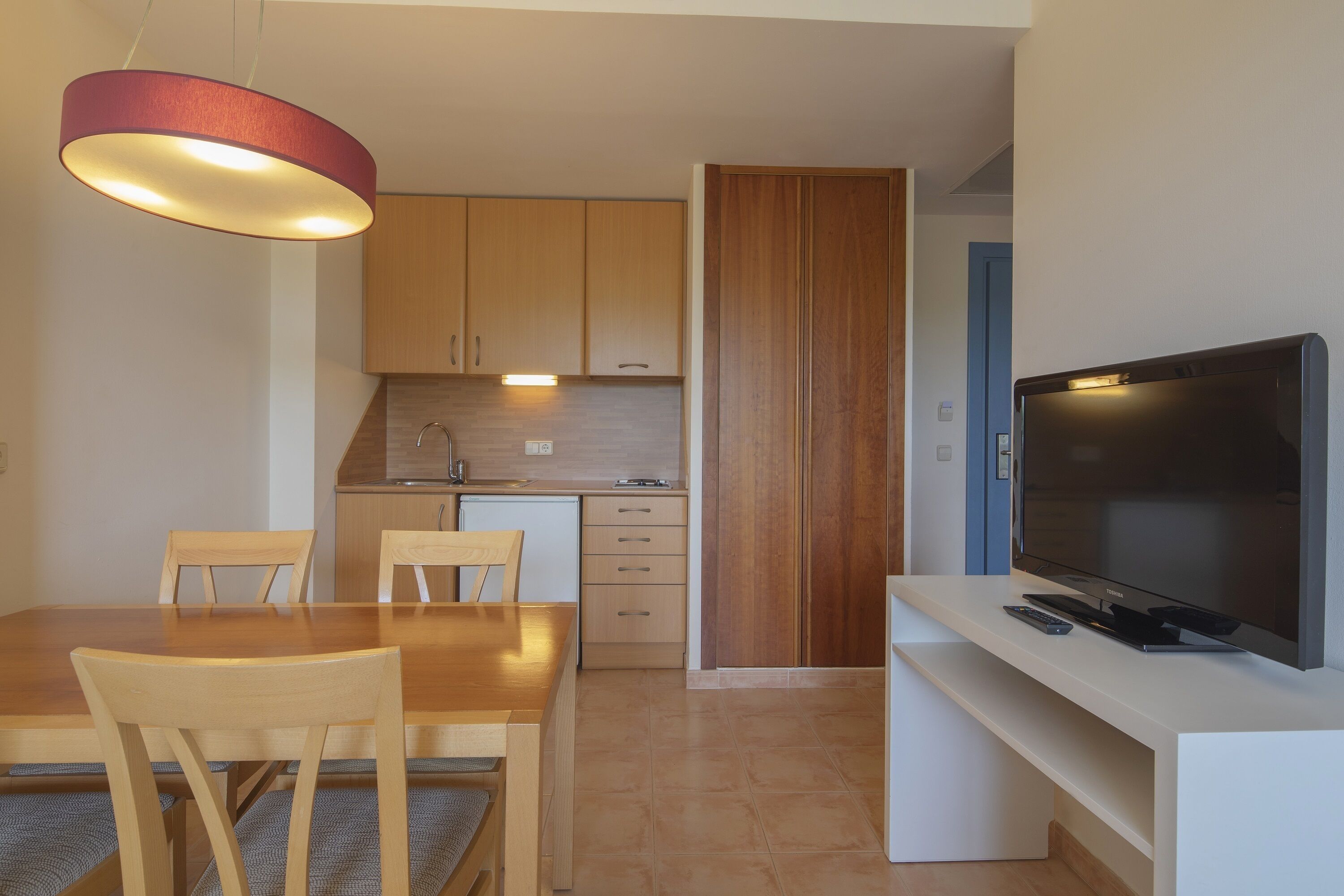 Een moderne keuken met houten kastjes, een eettafel met stoelen en een flatscreen televisie op een witte standaard.
