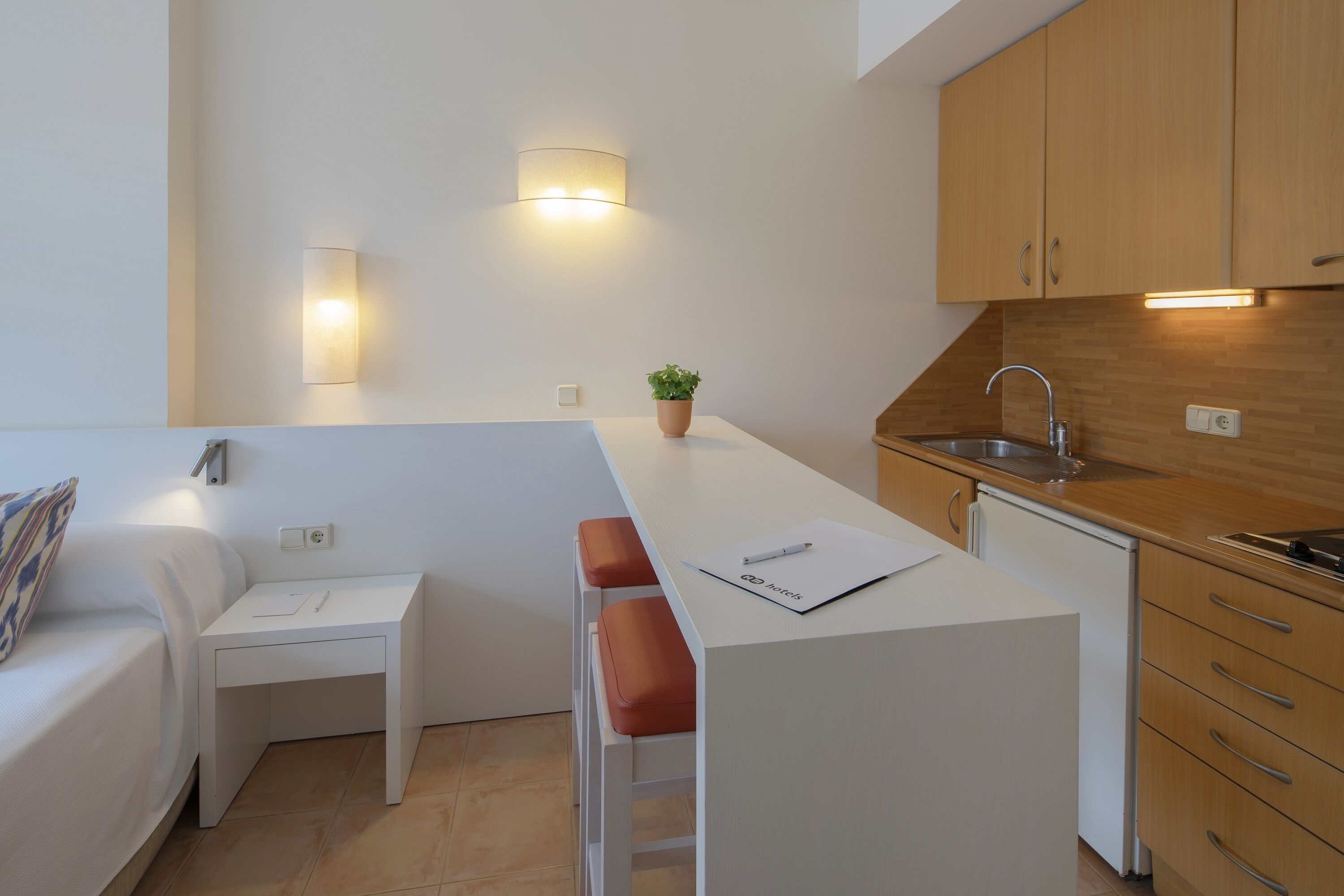 Cocina compacta con encimera blanca, armarios de madera, un lavamanos y una pequeña isla con asientos.