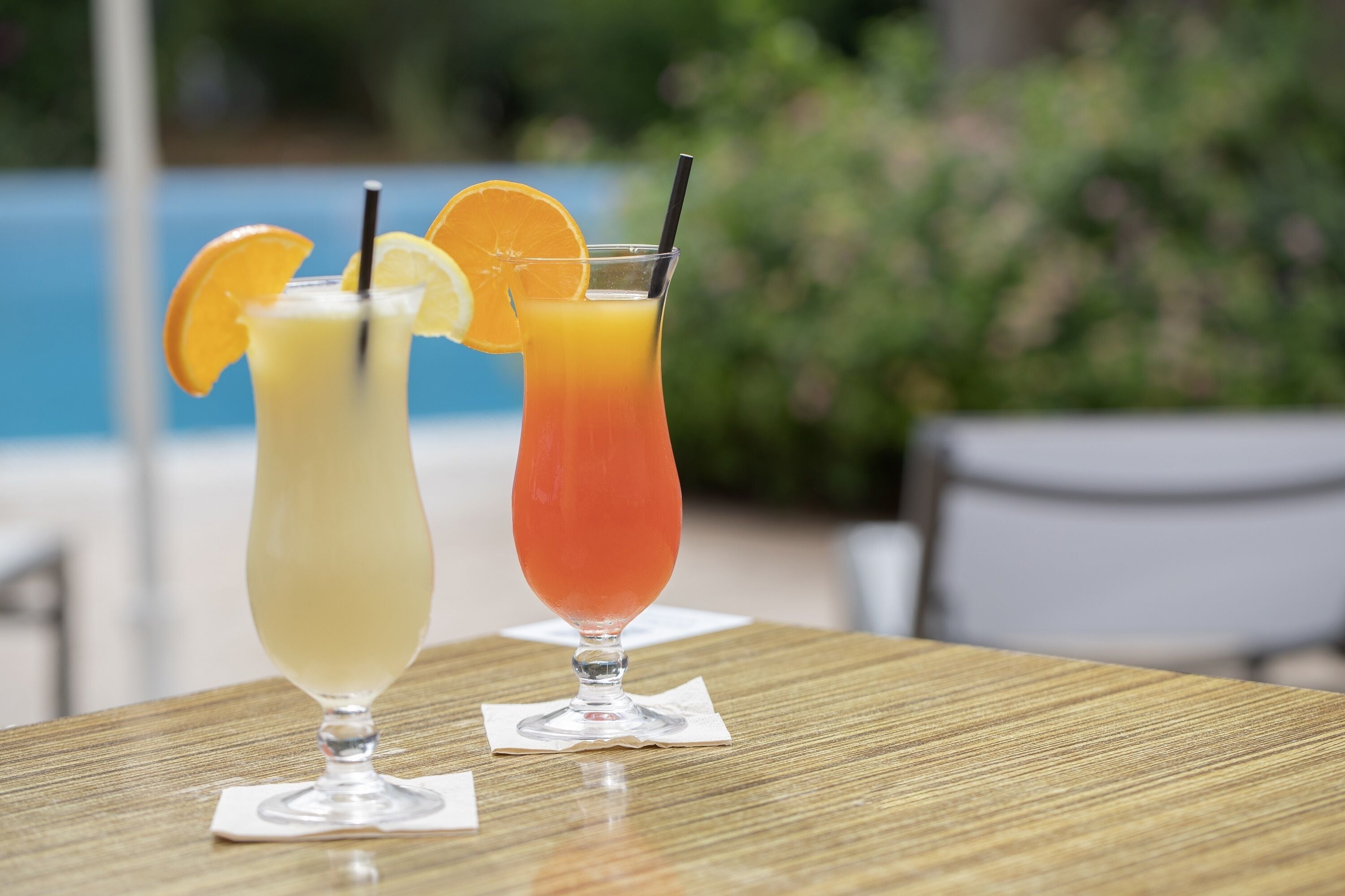 Two cocktails on a wooden table with a pool and greenery in the background.