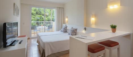 A hotel room with a bed, a desk with a TV, a small kitchen area, and a balcony with a view of greenery.