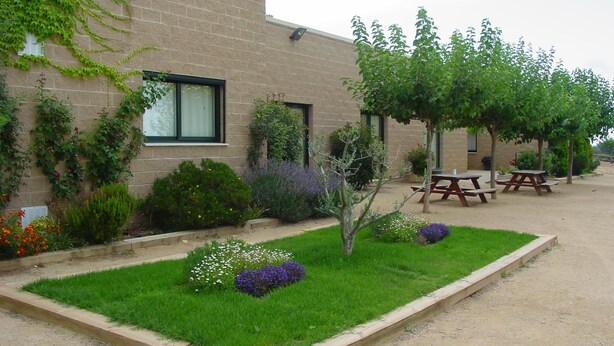 Edificio de ladrillo con jardín que incluye césped, flores y árboles. Se observan mesas de picnic y un banco.