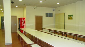 Un salón de conferencias con mesas largas y bancos, una máquina expendedora de Coca-Cola y una puerta.