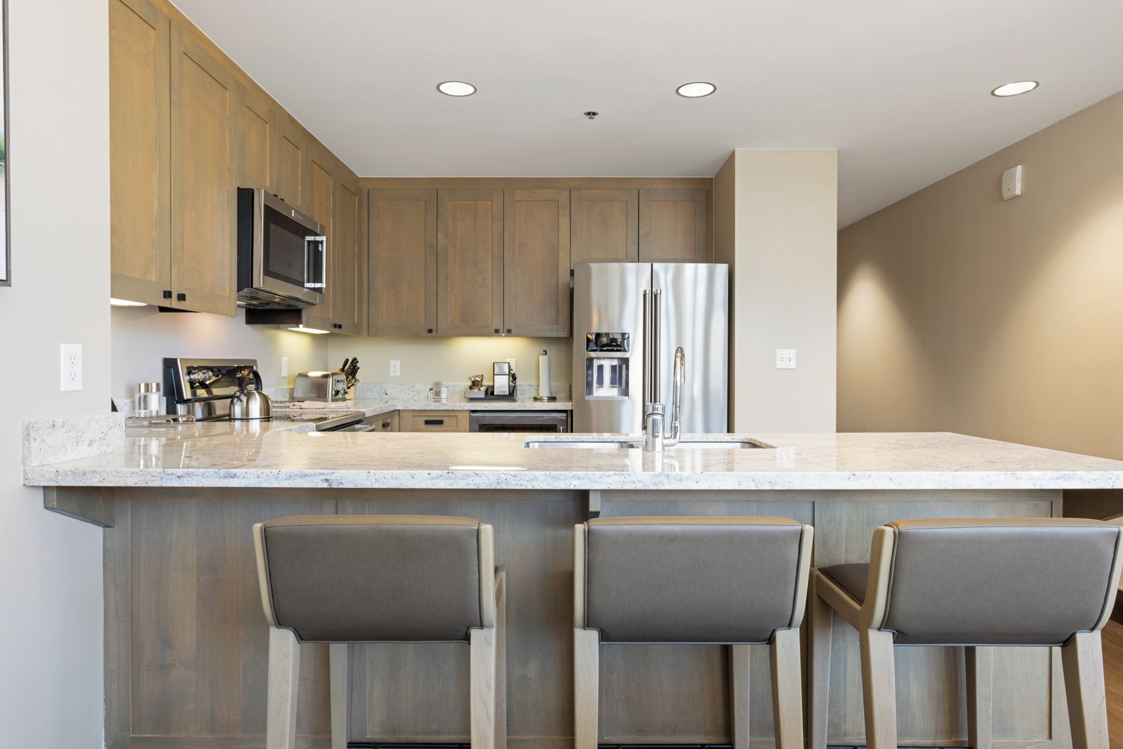 Una cocina moderna con armarios de madera, electrodomésticos de acero inoxidable y encimera de mármol. Hay tres taburetes de bar con asientos acolchados.
